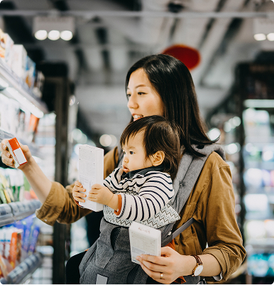 Mother holding her toddler in a carrier while shopping and examining products on a store shelf.