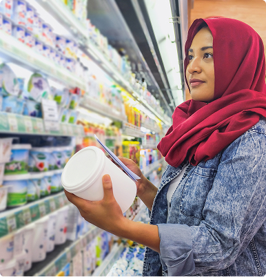 Woman in a red hijab and denim jacket reading product label in a grocery store dairy aisle.