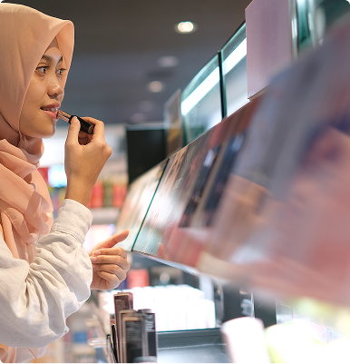 Woman in a peach-colored hijab applying lipstick in front of a store display mirror.