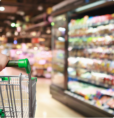 Hand pushing a shopping cart in a grocery store aisle with refrigerated shelves.