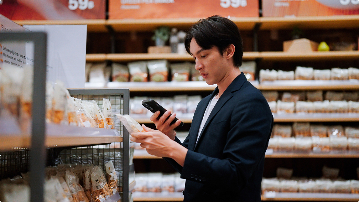 Young man in a dark blazer reading product information in a packaged food aisle while holding a smartphone.