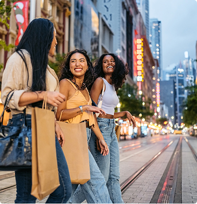 Three women smiling and walking with shopping bags on a city street at dusk.
