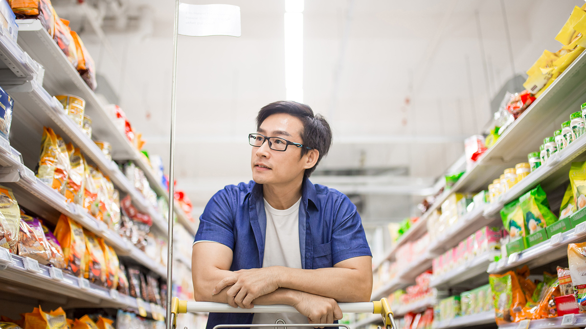 Man with glasses leaning on a shopping cart, looking at products in a brightly lit supermarket aisle.