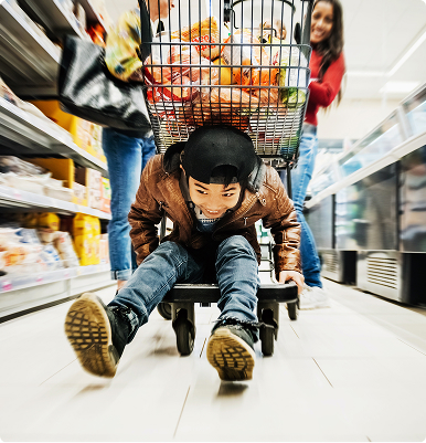 Child wearing a helmet sitting inside a shopping cart being pushed down a grocery store aisle by a smiling adult.