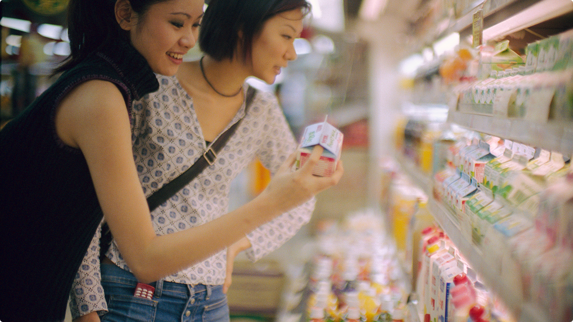 Two women smiling and examining a carton of beverage in a brightly lit supermarket aisle.