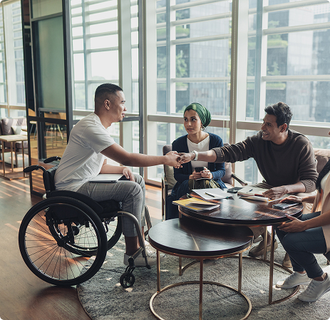 A man in a wheelchair shakes hands with a seated man at a round table while a woman in a hijab watches attentively, in a modern office setting.