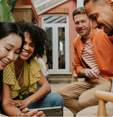 Four friends sitting closely, smiling and looking at a smartphone together indoors.