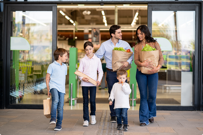 Family of five leaving a grocery store carrying paper bags filled with fresh produce.