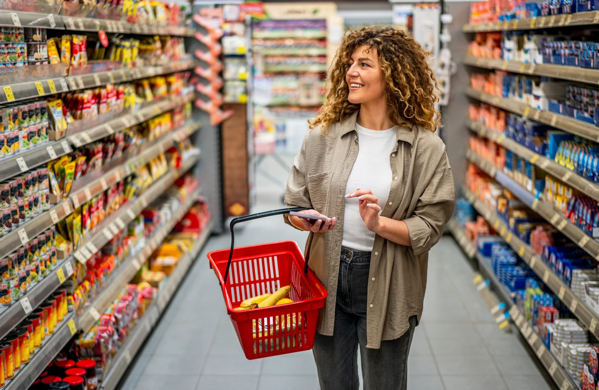 Smiling woman holding a red shopping basket with bananas and a phone while standing in a grocery store aisle.