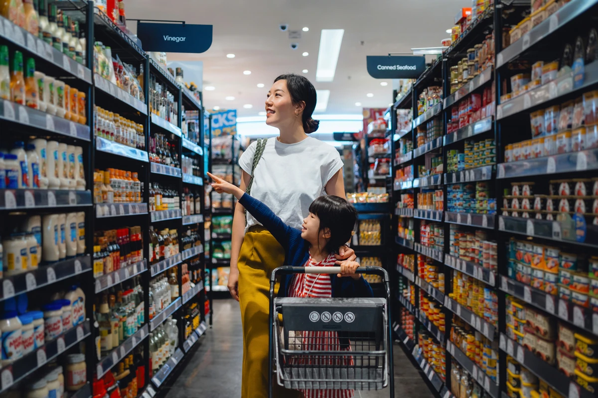 Mother and daughter shopping in a grocery aisle with shelves stocked with vinegar and canned seafood.