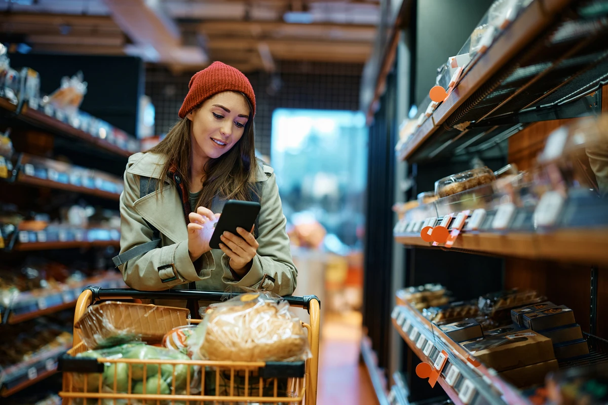 Woman in a red beanie and coat shopping with a cart full of groceries while using a smartphone in a grocery store aisle.