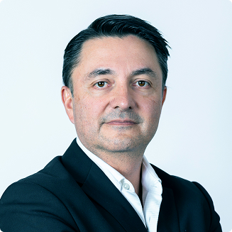 Professional man with dark hair wearing a white collared shirt and black blazer against a light background.