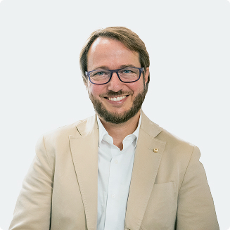Smiling man with glasses, beard, and light brown blazer over white shirt against a plain background.