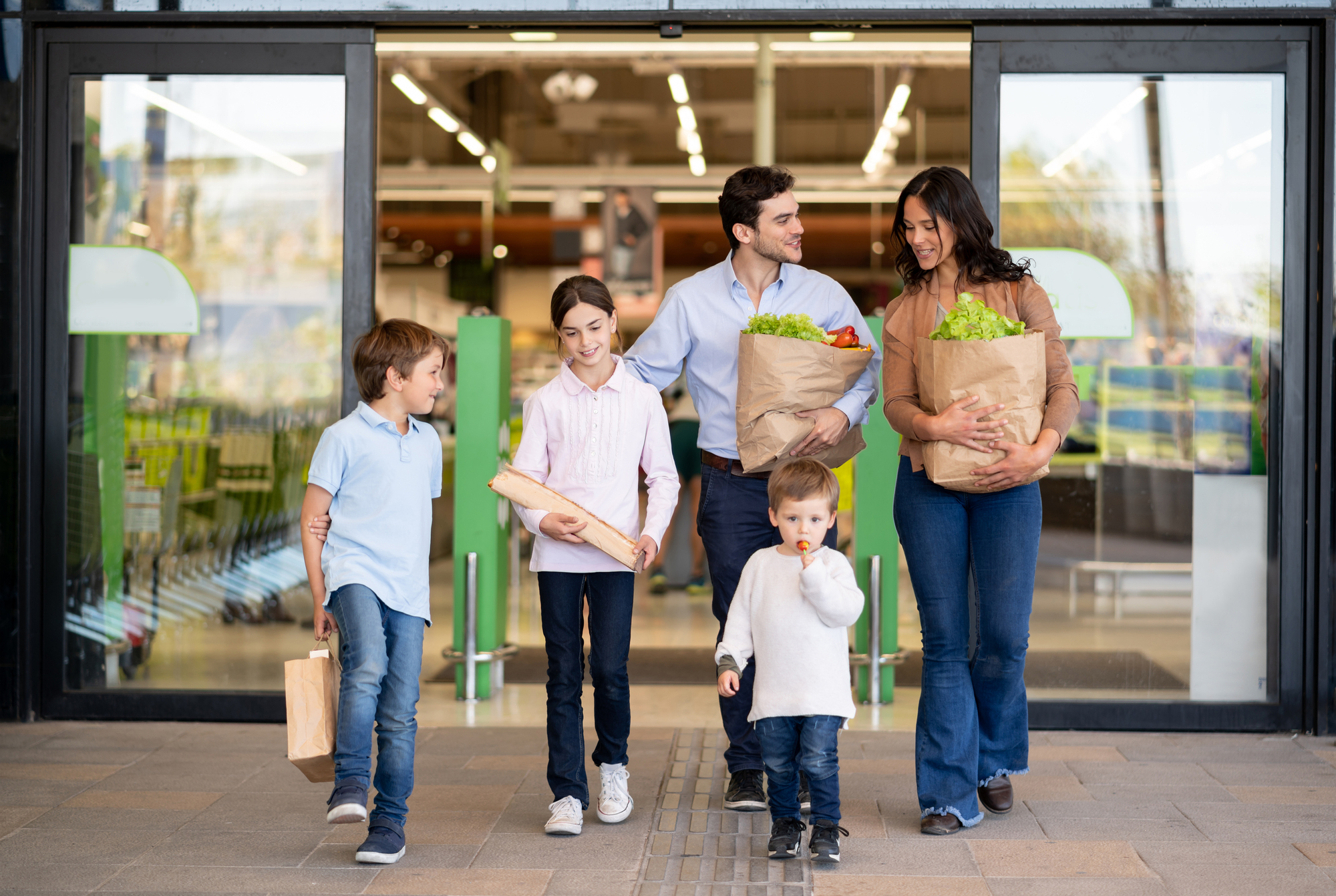 A family of two adults and three children walking out of a grocery store with paper bags of groceries and a baguette.