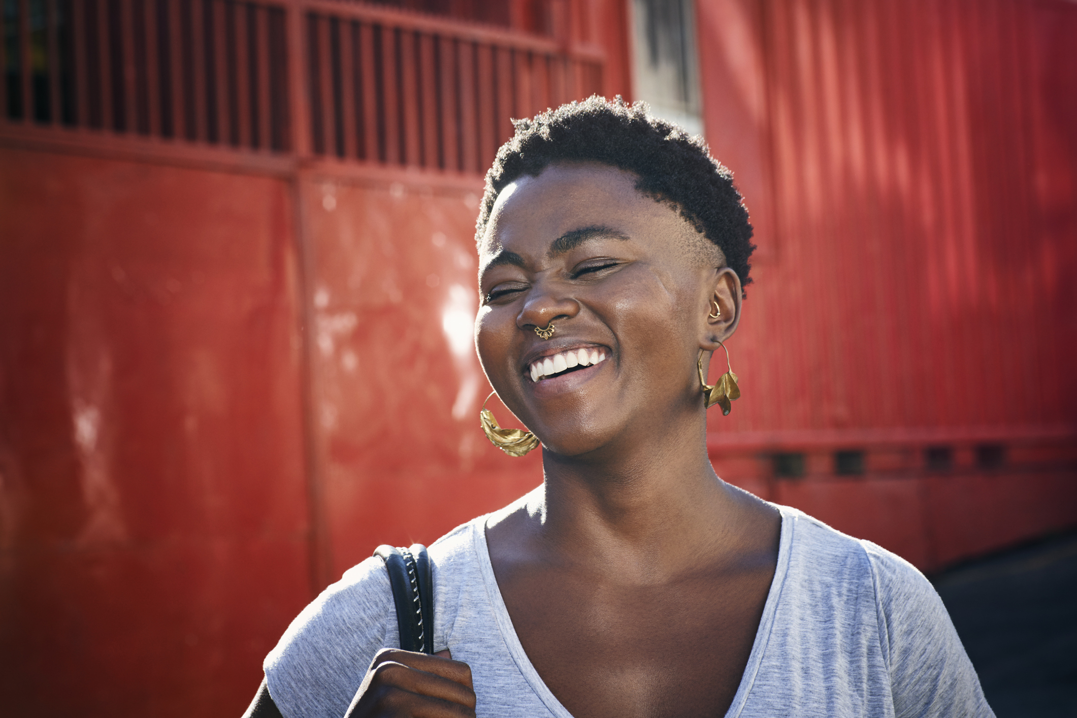 Smiling young woman with short curly hair, gold earrings, and a septum ring standing in front of a red wall.