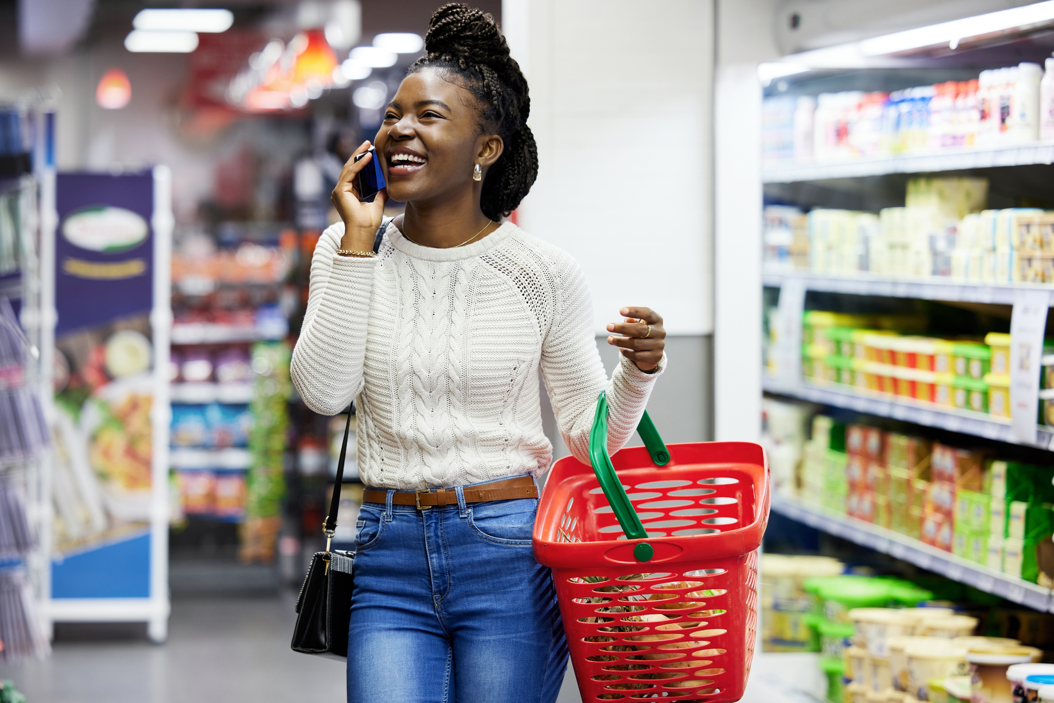 Smiling woman holding a red shopping basket and talking on a phone in a grocery store aisle.
