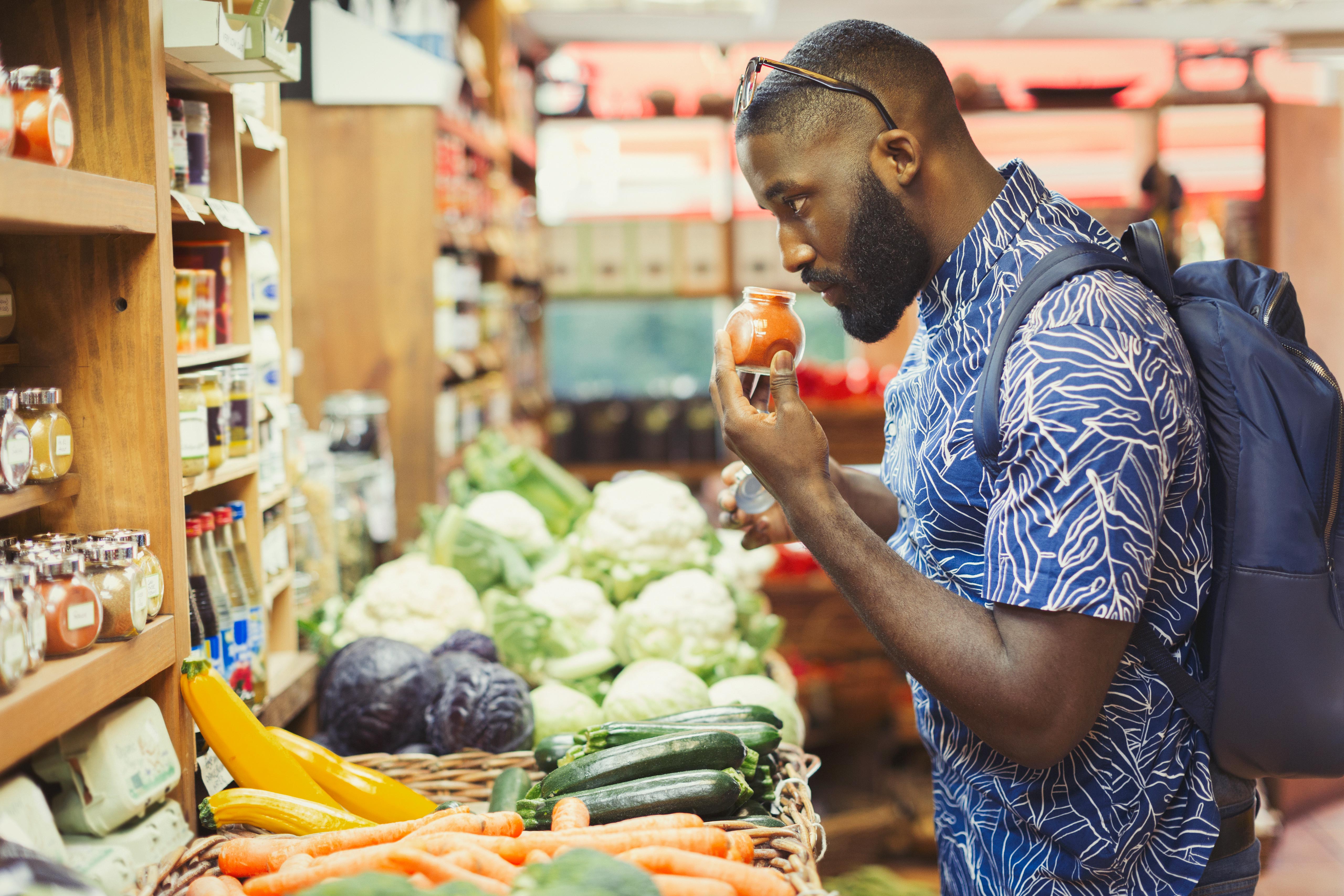 Man in blue patterned shirt smelling a spice jar while shopping for vegetables in a market.