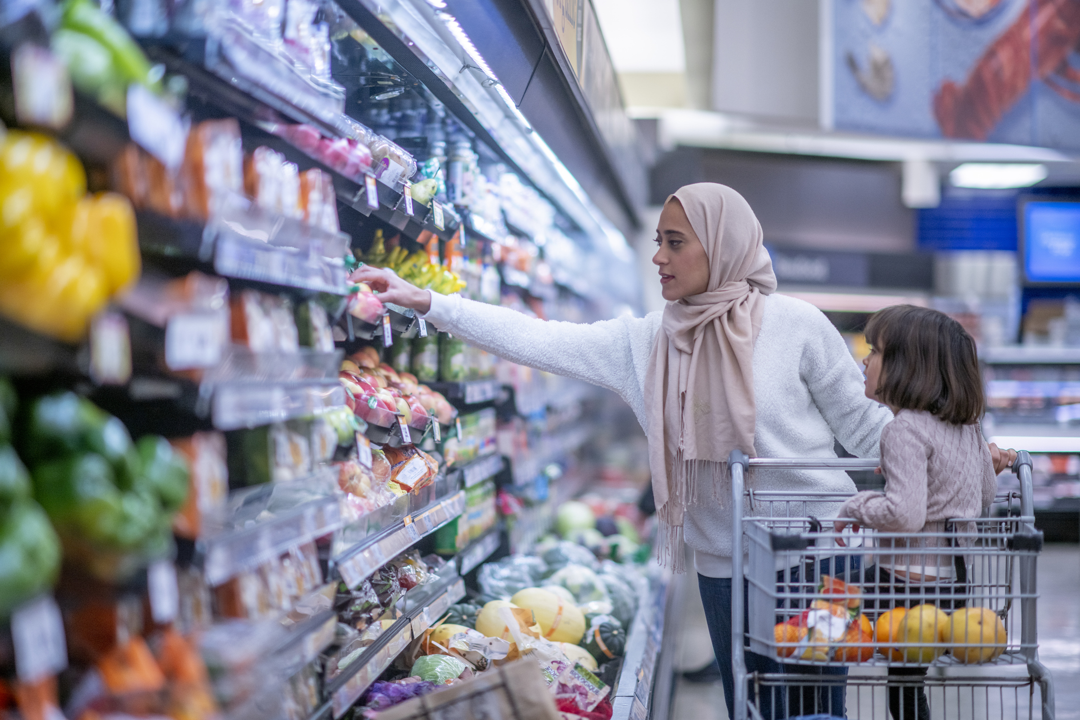 Woman wearing a hijab reaching for produce on a grocery store shelf while a child sits in the shopping cart filled with fruits and vegetables.