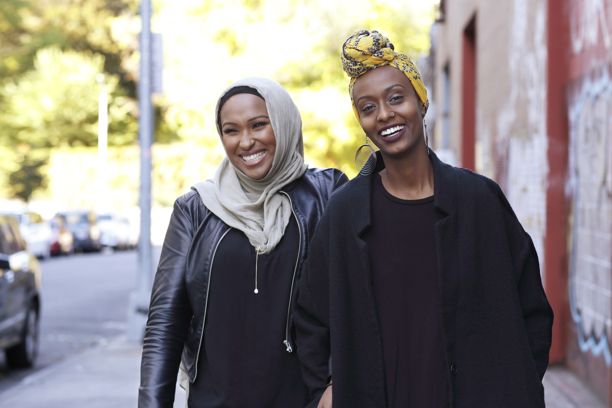 Two smiling women walking outdoors on a city street, one wearing a light-colored hijab and leather jacket, the other wearing a yellow patterned headwrap and black coat.