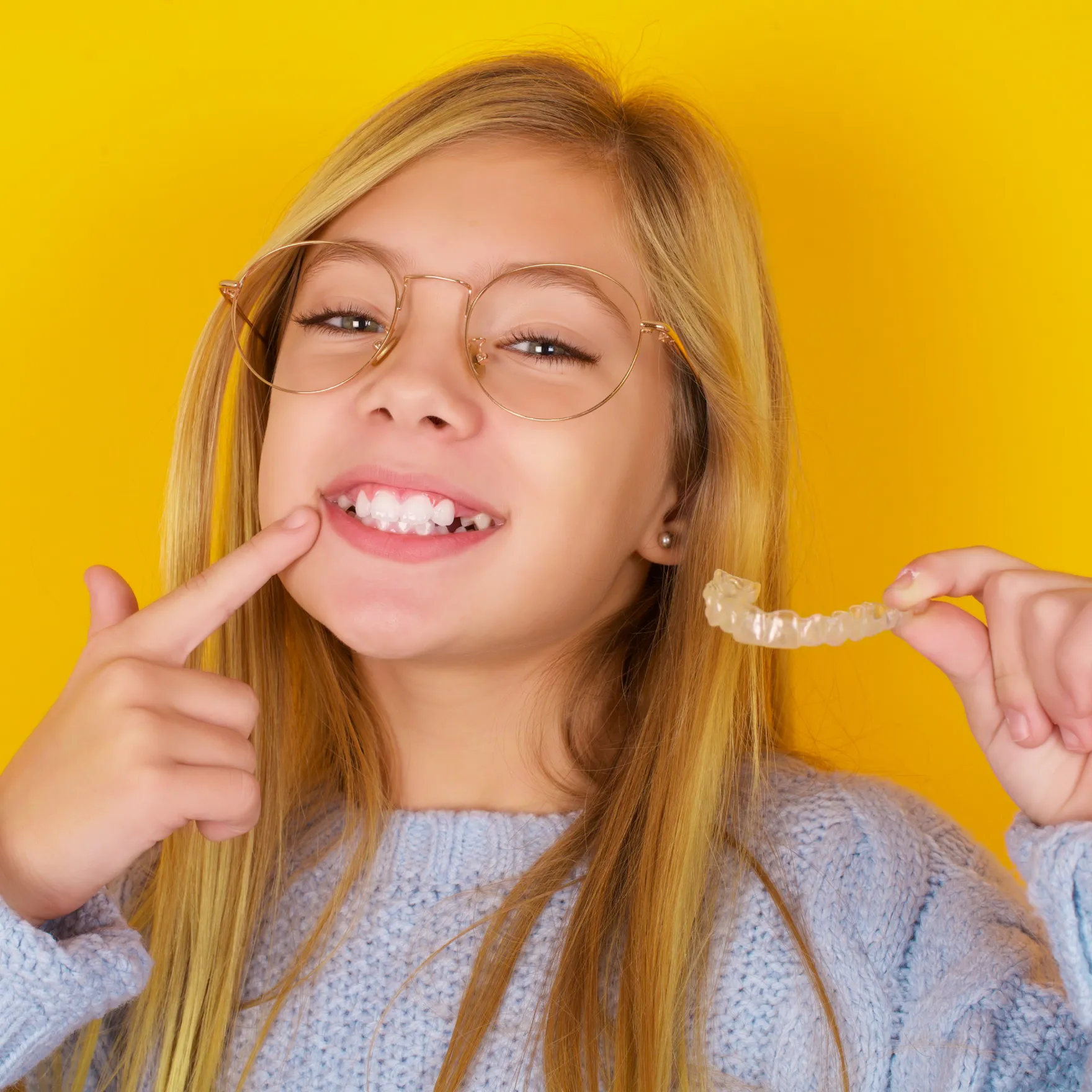 Invisalign in New York, NY | girl smiling holding an Invisalign Tray and pointing at her smile