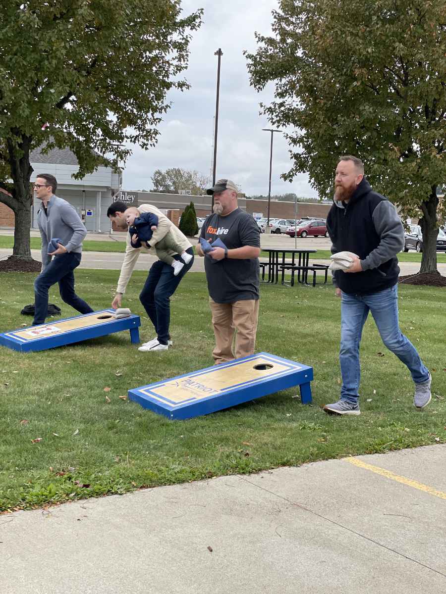 Four men playing cornhole on a grassy area with trees and buildings