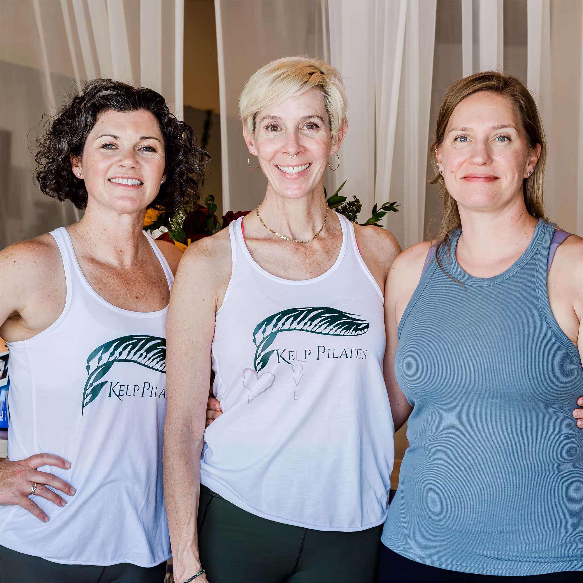 Three women smiling indoors wearing athletic tops, two in white tank tops with Kelp Pilates logos.