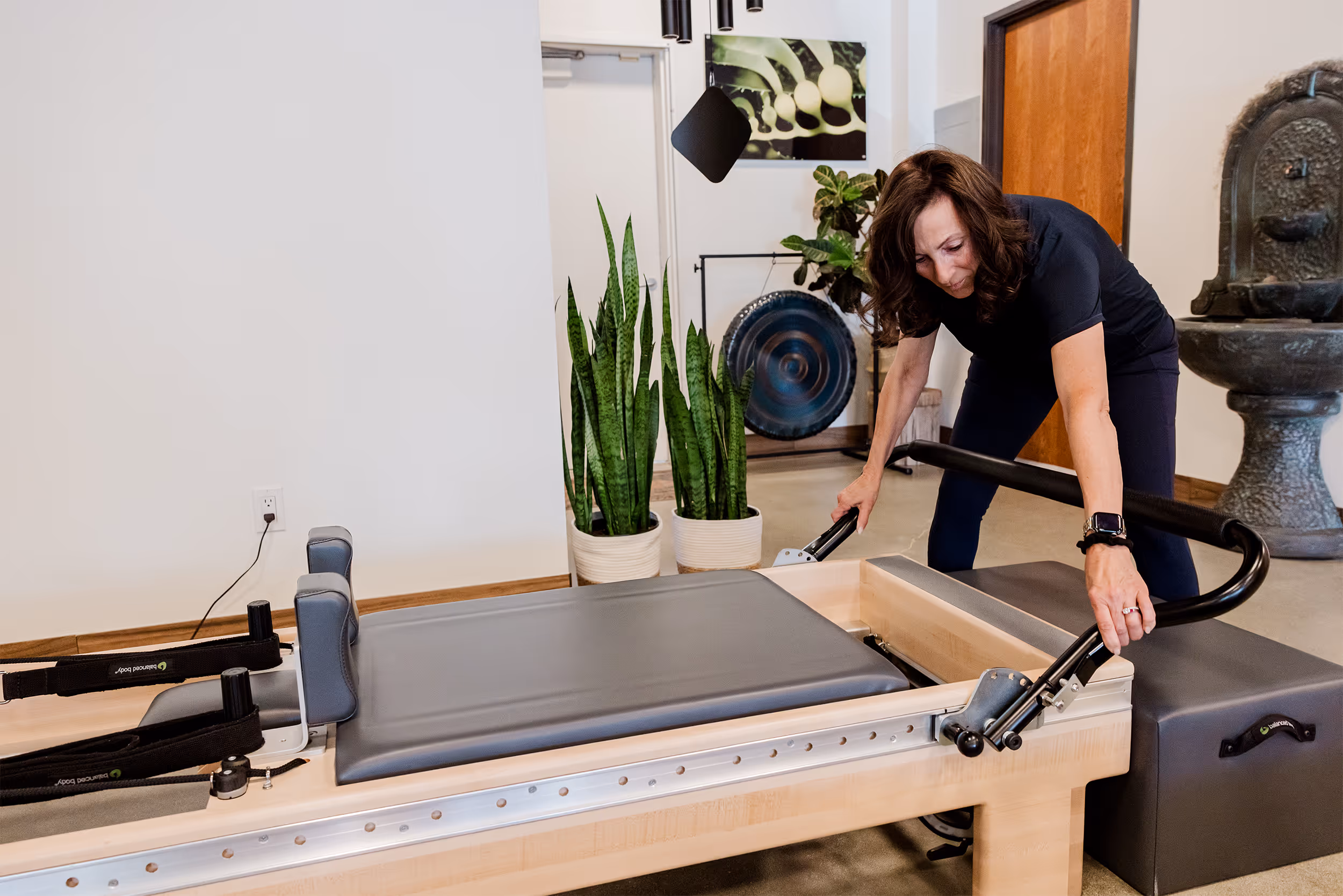 Woman adjusting a reformer Pilates machine in a modern studio with plants and a gong in the background.