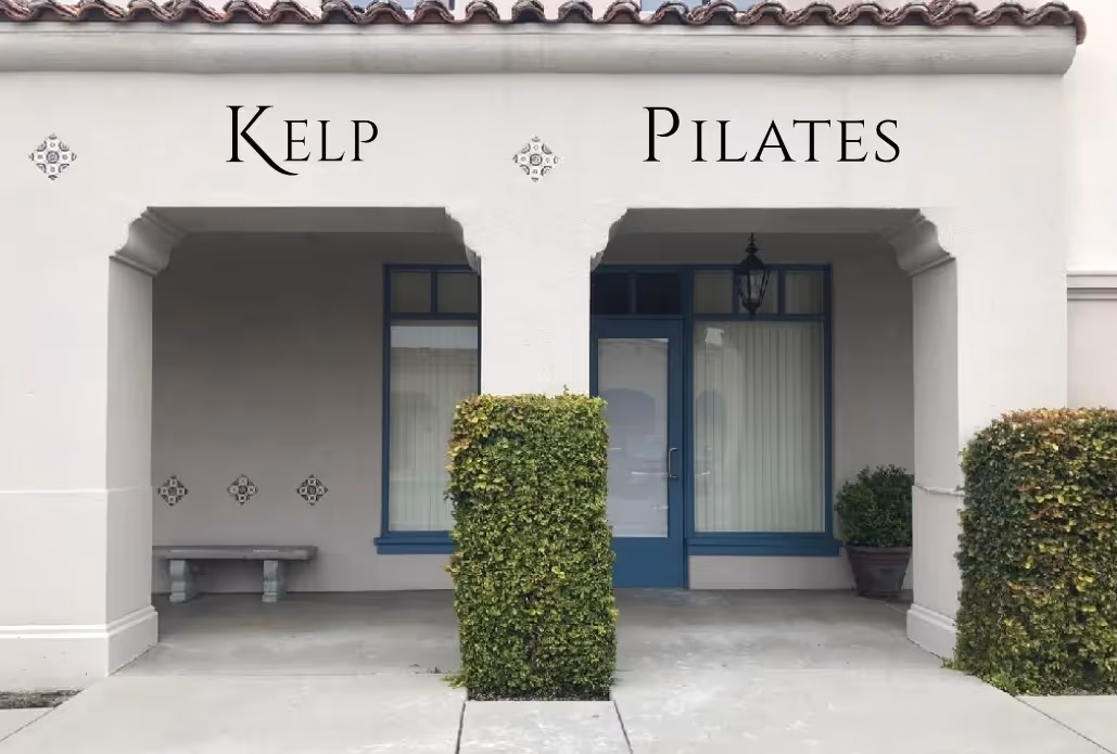 Entrance of a building with two archways labeled Kelp and Pilates, featuring a bench, planter, and neatly trimmed shrubs.