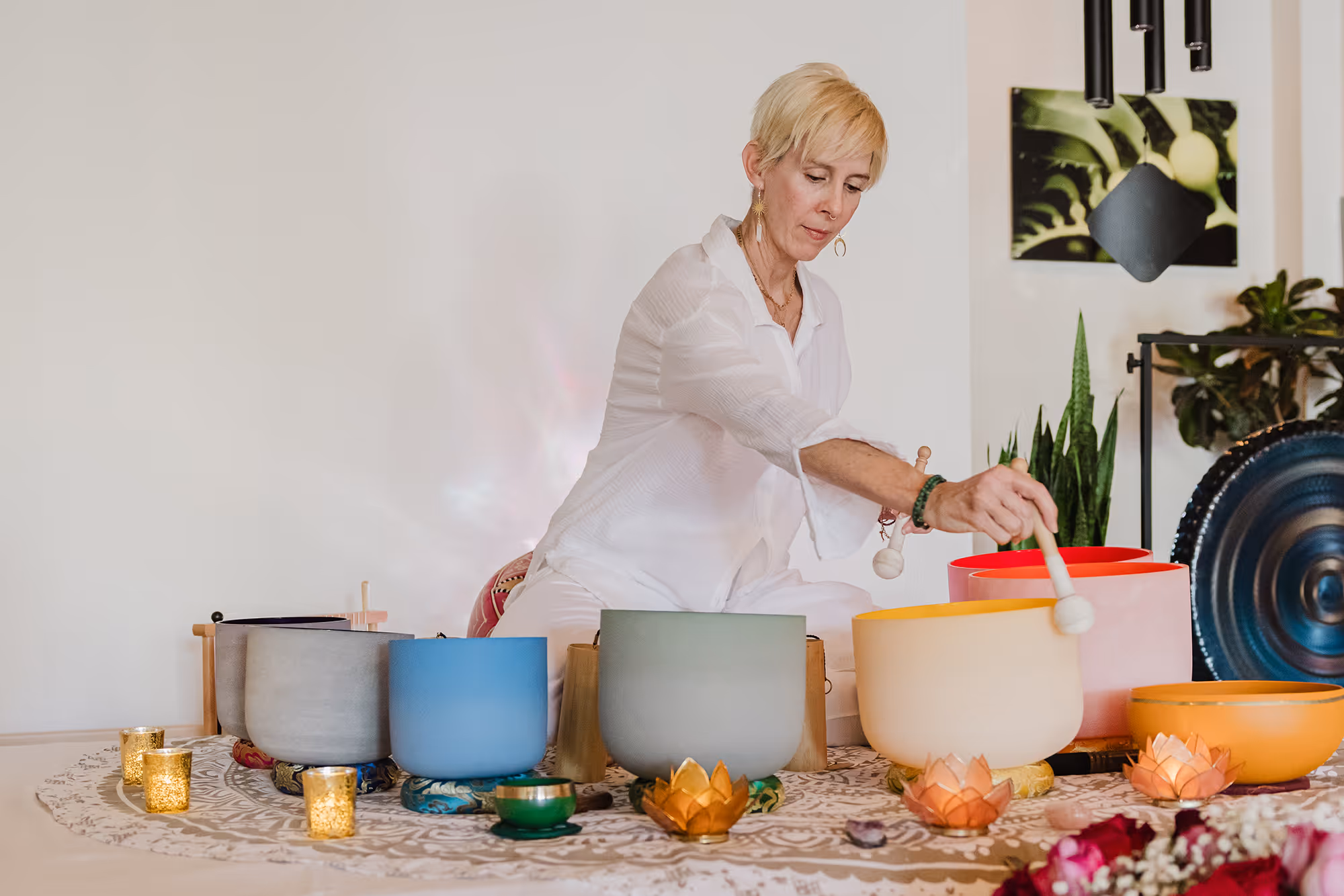 Woman in white clothes playing multicolored crystal singing bowls arranged on a patterned cloth with candles and plants in the background.