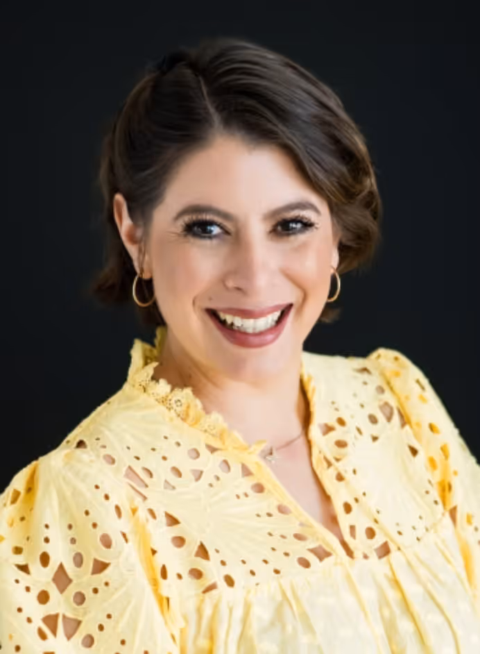 Smiling woman with short brown hair wearing a yellow eyelet dress and gold hoop earrings against a black background.