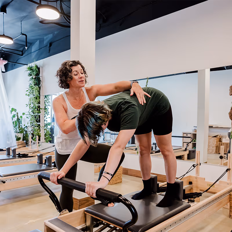 Pilates instructor helps a student perform a stretch on a reformer machine in a bright studio.