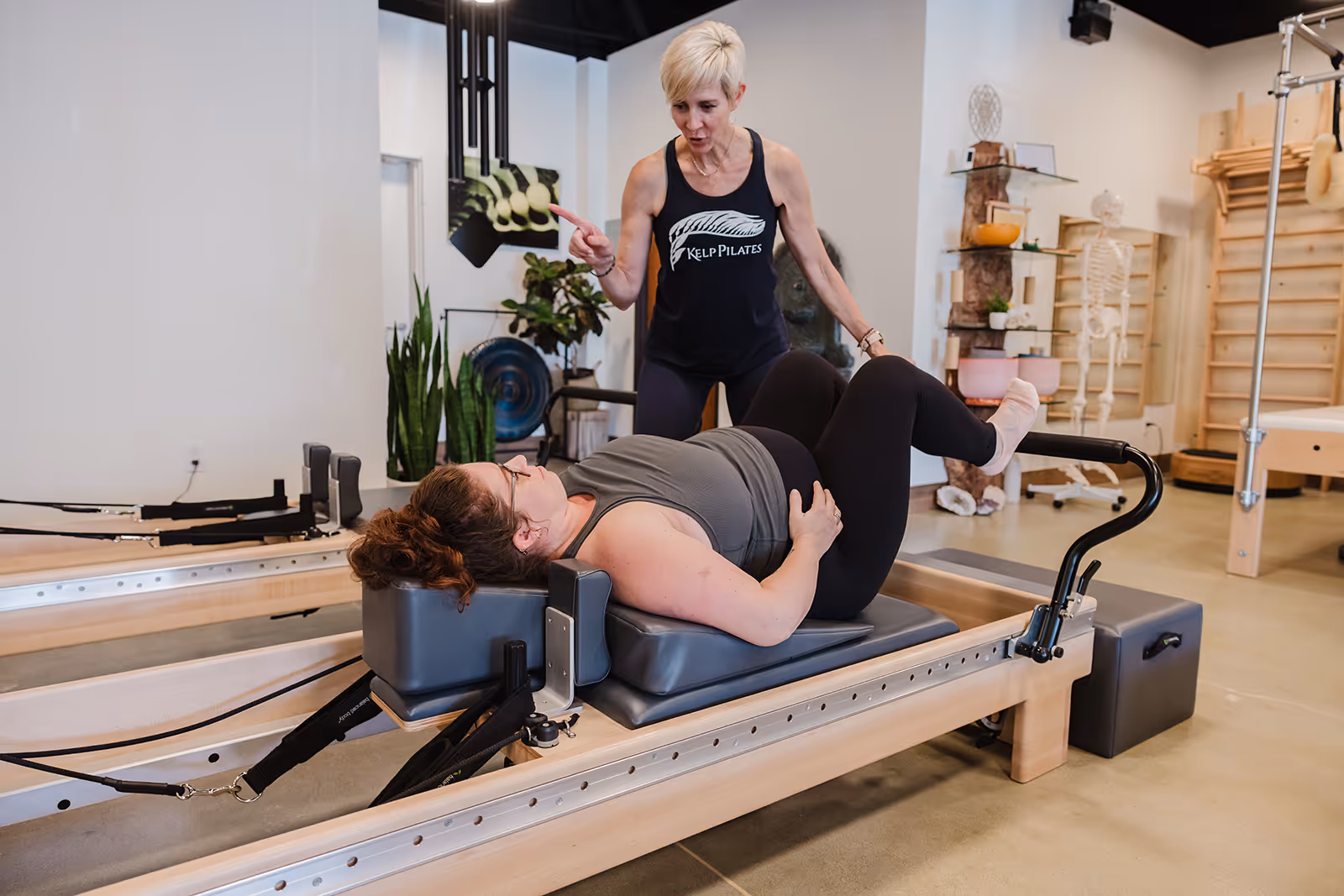 Woman lies on Pilates reformer machine with knees bent while instructor in Kelp Pilates tank top coaches her.