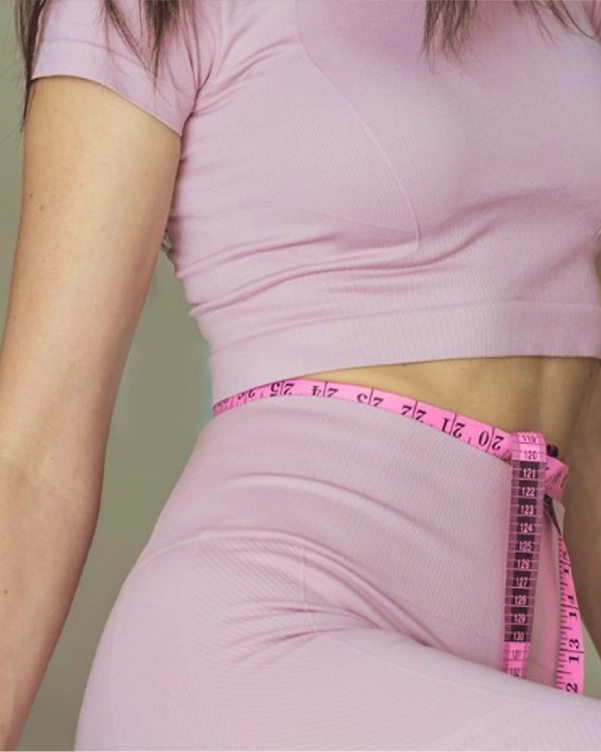 O femeie își măsoară talia cu o bandă de măsurare roz, evidențiind concentrarea pe fitness sau controlul greutății.