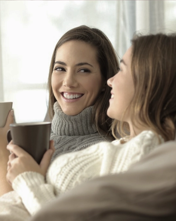Două femei zâmbitoare și relaxate stau pe o canapea cu cești de cafea sau ceai, bucurându-se de compania celeilalte.