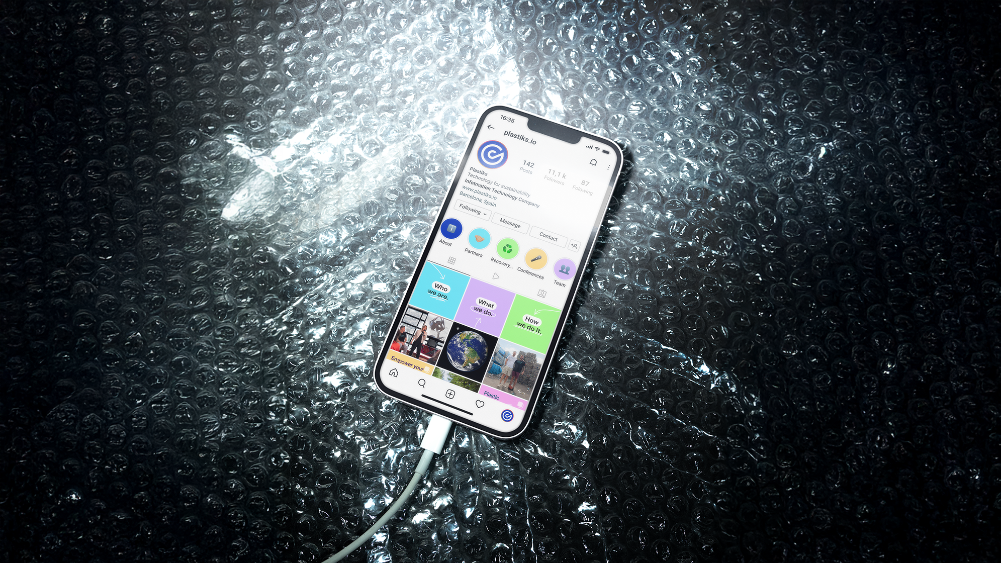 Smartphone displaying an Instagram profile about sustainability and technology, lying on black bubble wrap with a charging cable connected.