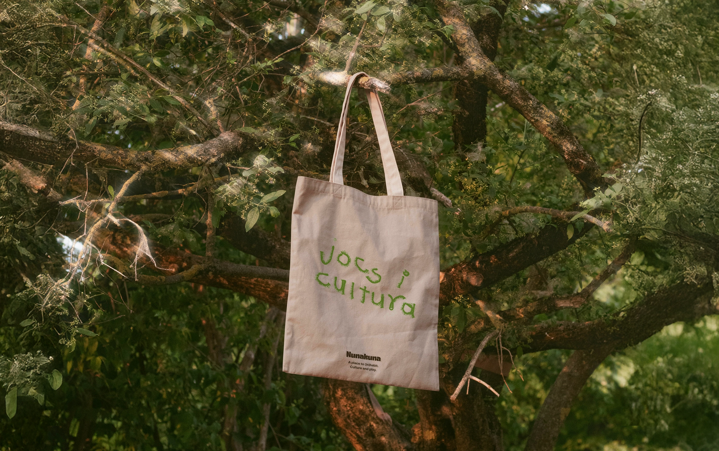 Cream tote bag with green text 'jocs i cultura' and black text 'Nunakuna A place to (in)habit. Culture and play.' hanging from a tree branch in a leafy outdoor setting.