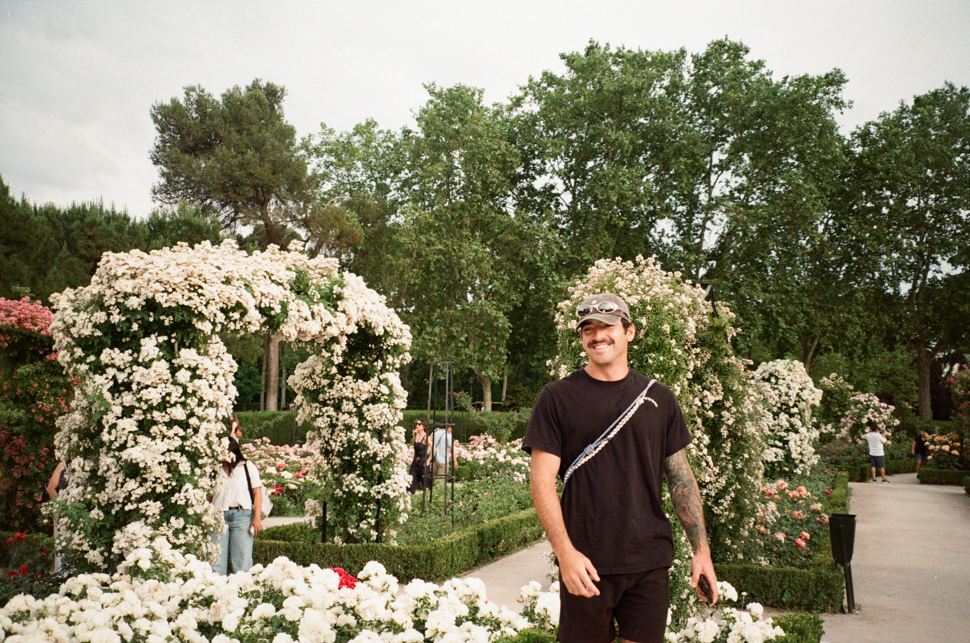 Man smiling and walking in a garden with white flowering arches and green trees in the background.