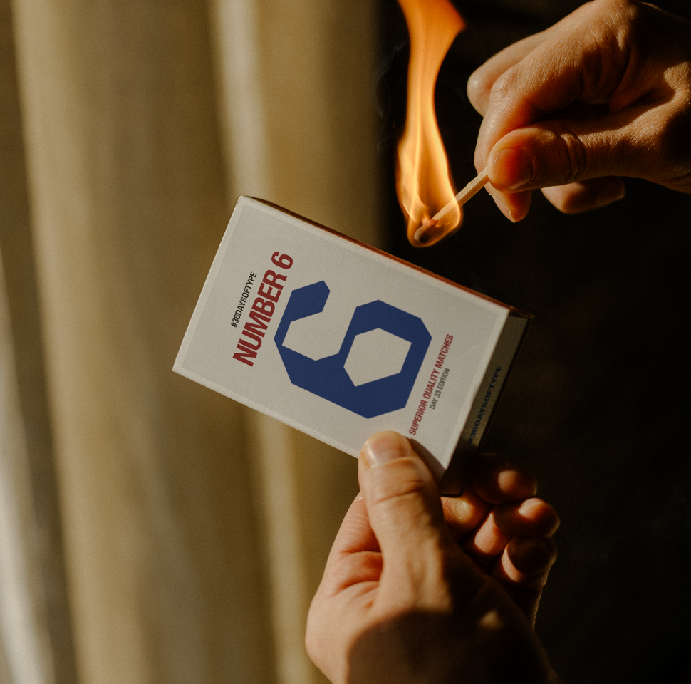 Hands holding a matchbox labeled 'Number 6' and lighting a match with a bright flame.