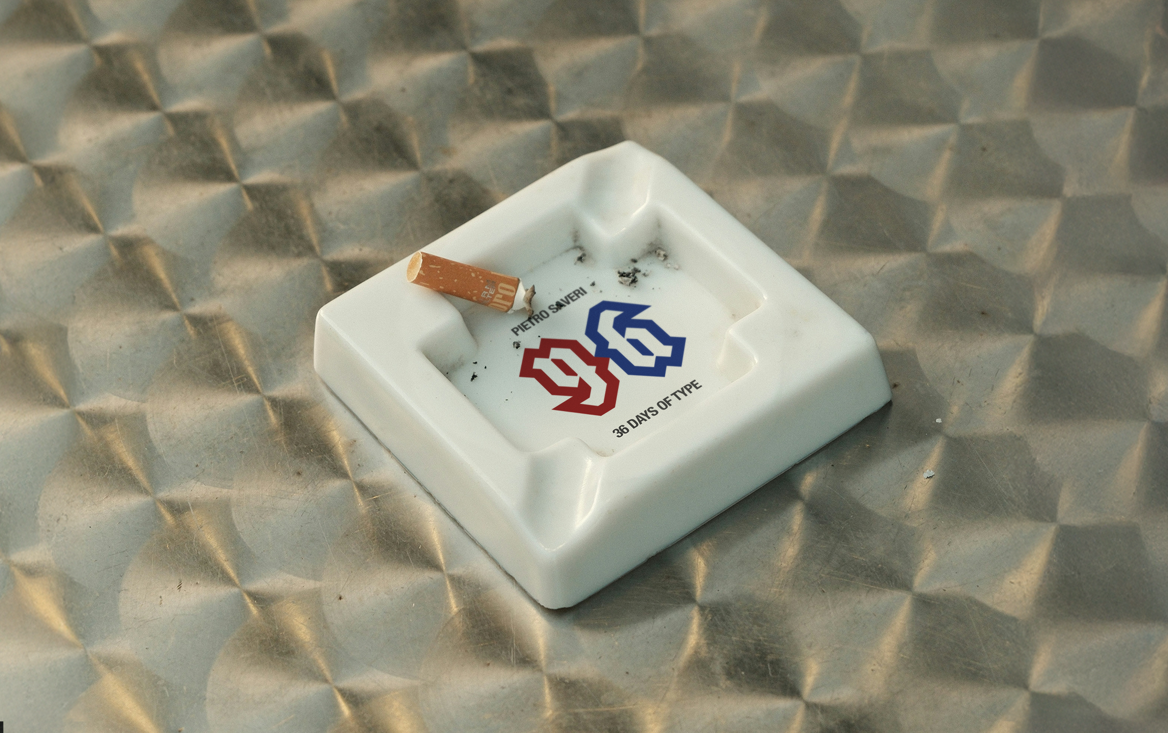 White square ashtray with a red and blue 36 Days of Type logo and a cigarette butt on a metallic textured surface.