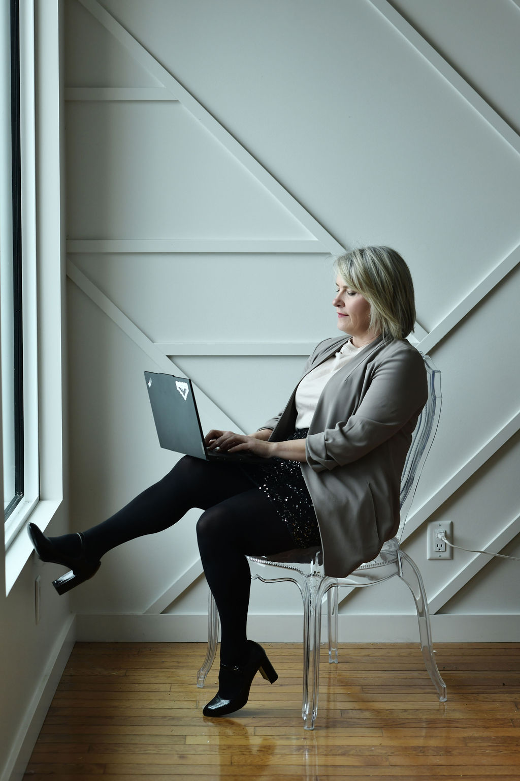blonde woman in grey top and black skirt and shoes, sits legs crossed on a chair working at a laptop