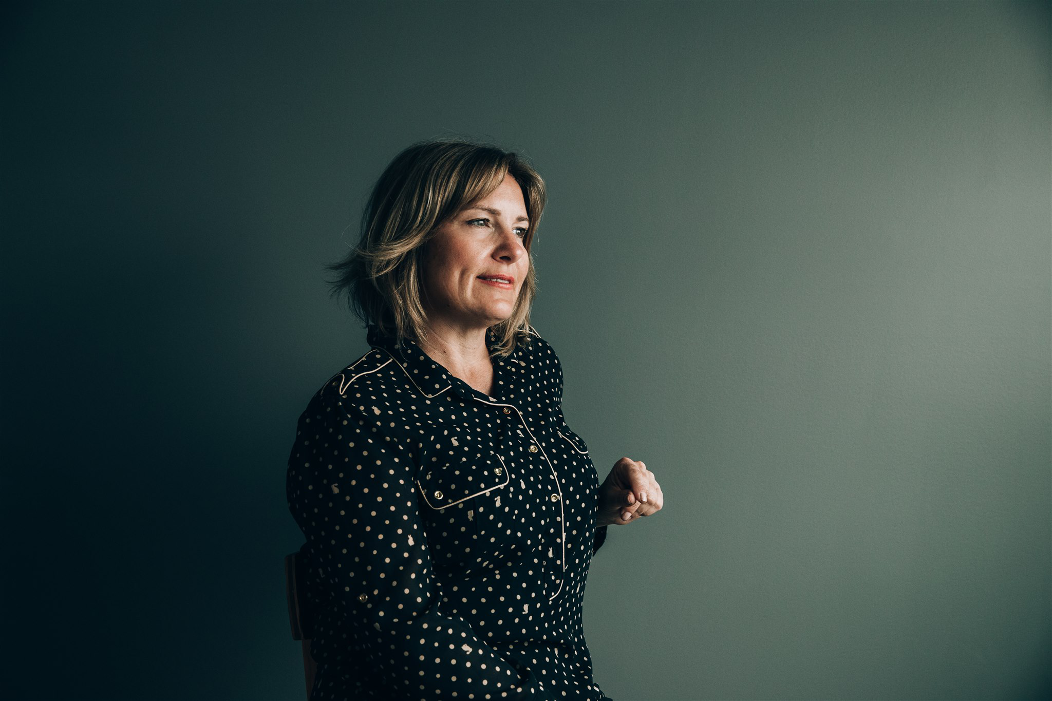 A woman in a black polka dot blouse sits against a dark green wall, her hand raised as if in conversation