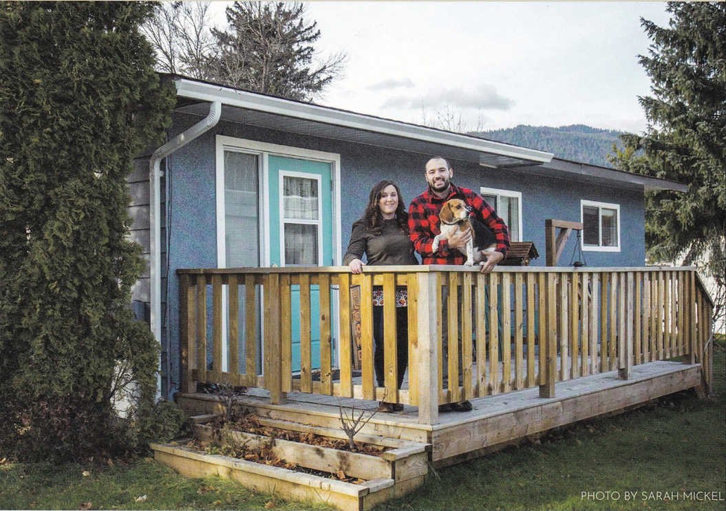 A couple stands on a wooden porch of a blue house, holding a small dog, surrounded by greenery and mountain views