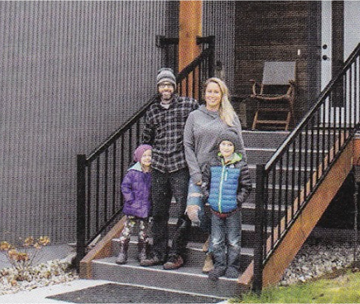 A family stands at the bottom of stairs, in-front of a grey house with white door
