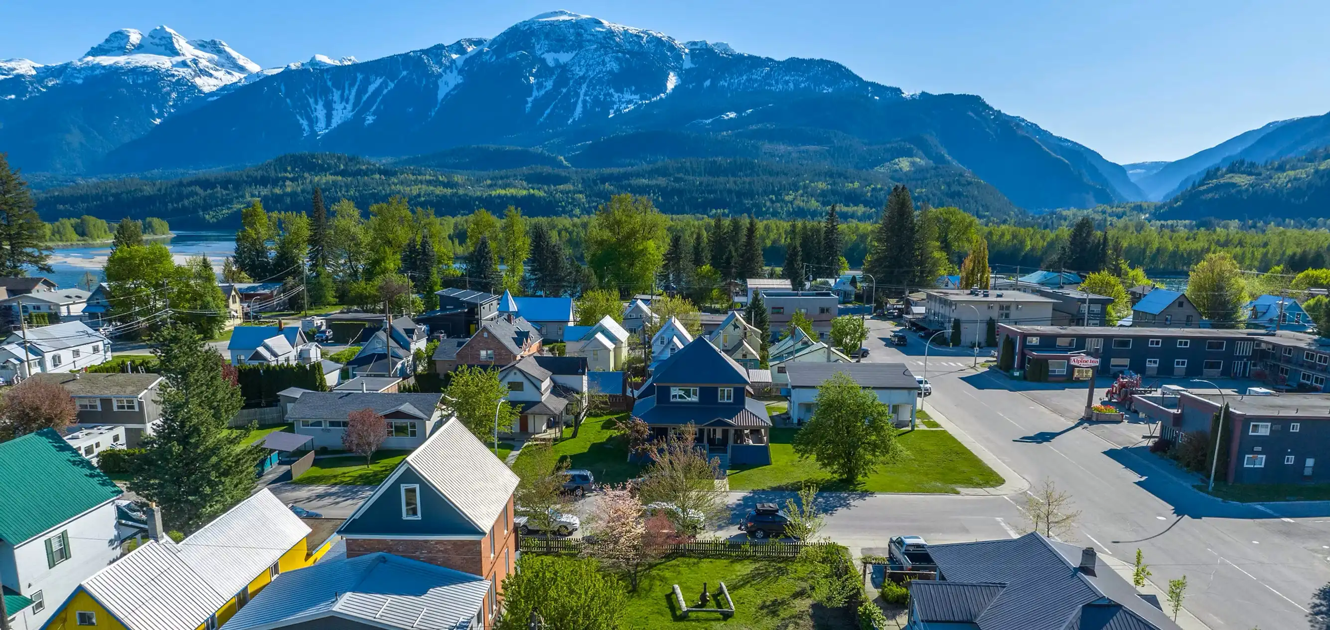 Aerial view of a Revelstoke town with colorful houses, lush greenery, and snow-capped mountains in the background on a clear day