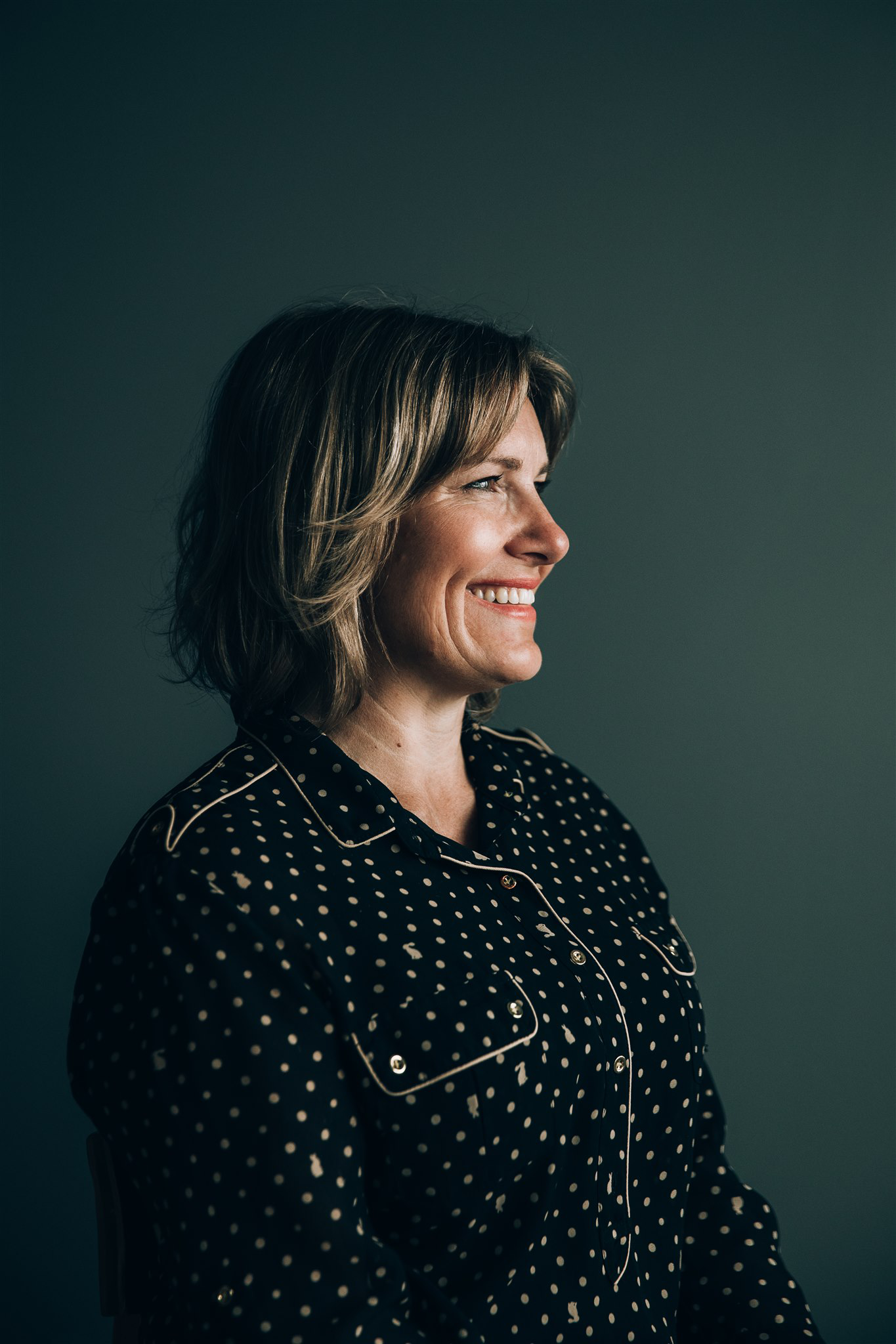 A smiling woman in a black polka dot blouse sits against a dark green wall