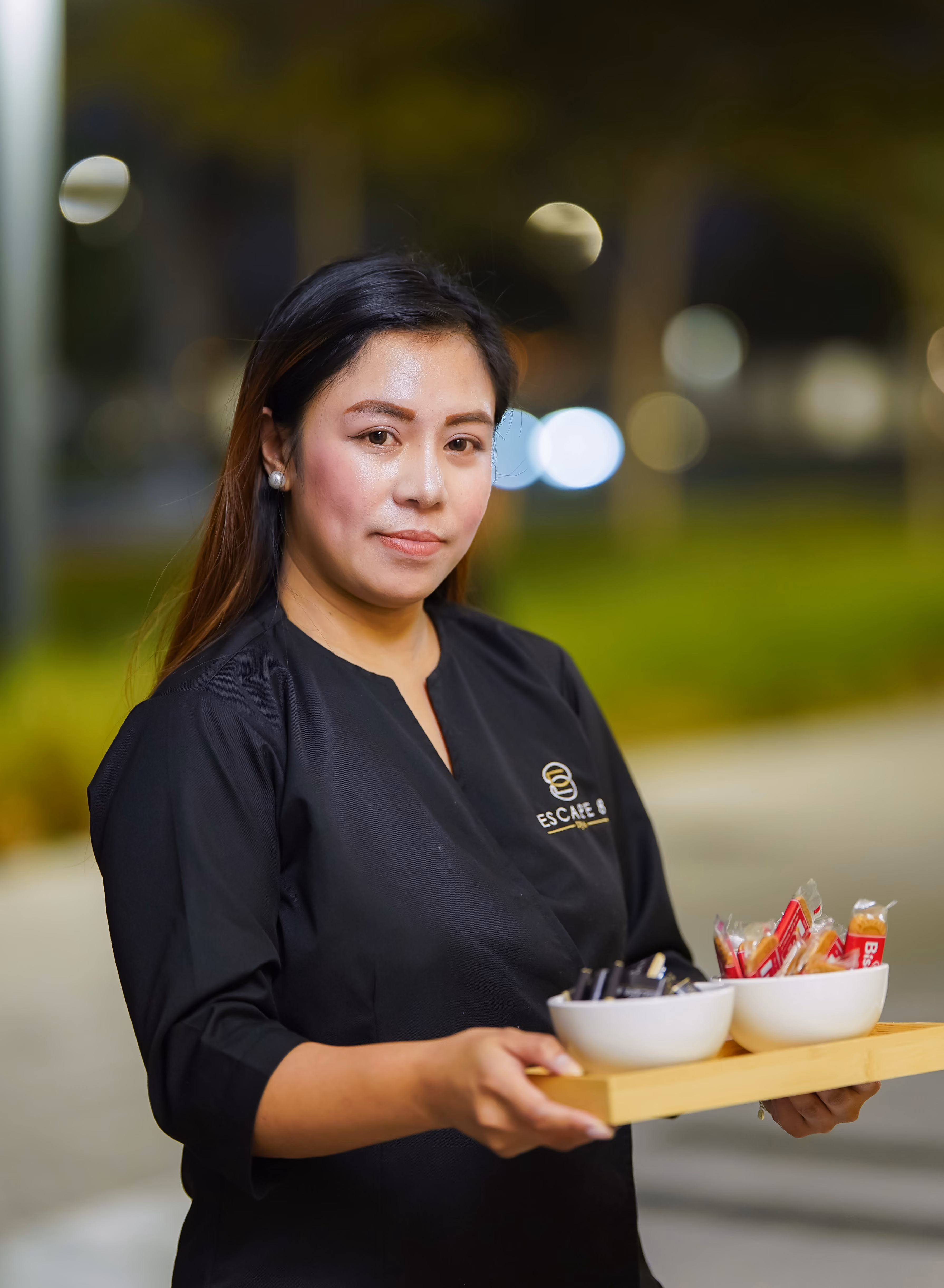 Woman in black uniform holding a wooden tray with two bowls of wrapped snacks outdoors at night.