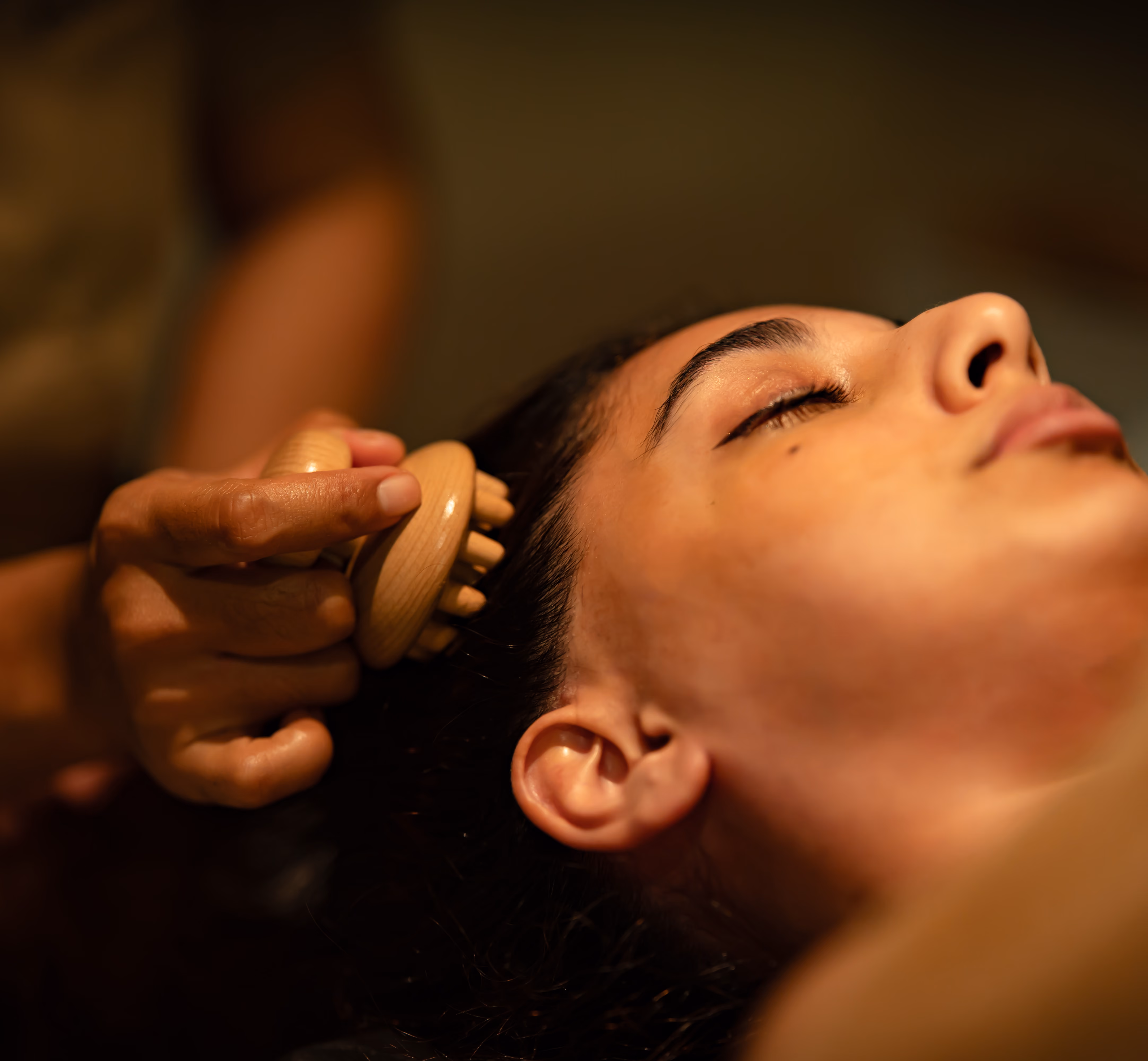 Person receiving a gentle scalp massage with a wooden massage tool on dark hair in warm lighting.