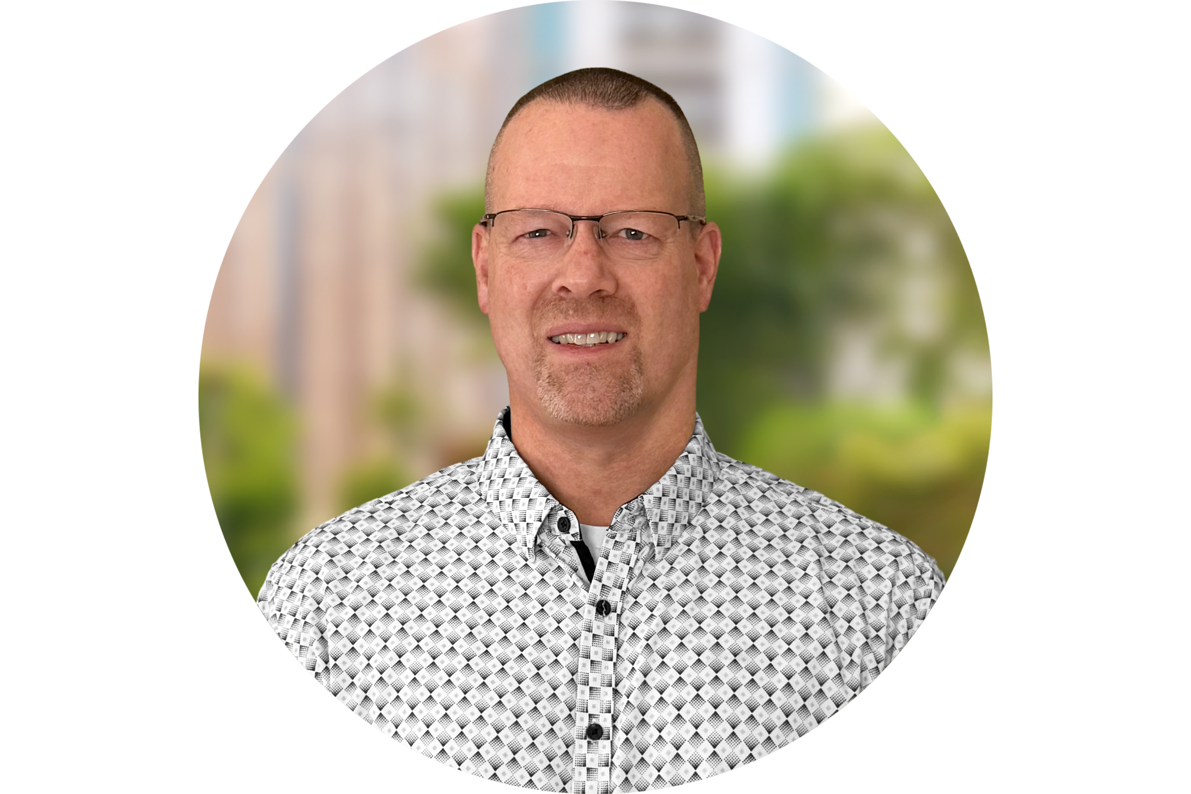 Man with short hair and glasses wearing a patterned button-up shirt, smiling slightly with a blurred outdoor background.