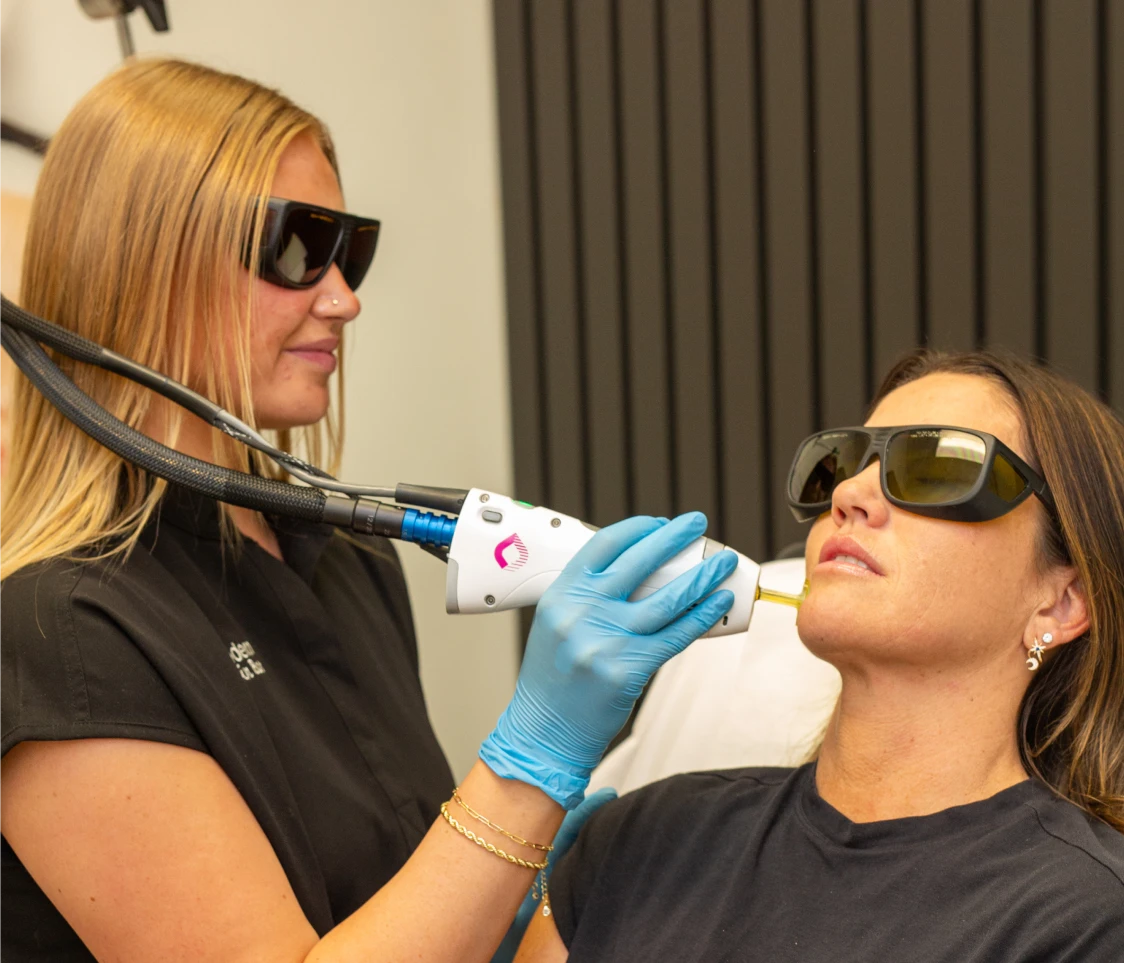 Woman wearing protective glasses receiving a laser treatment on her chin from a gloved technician.