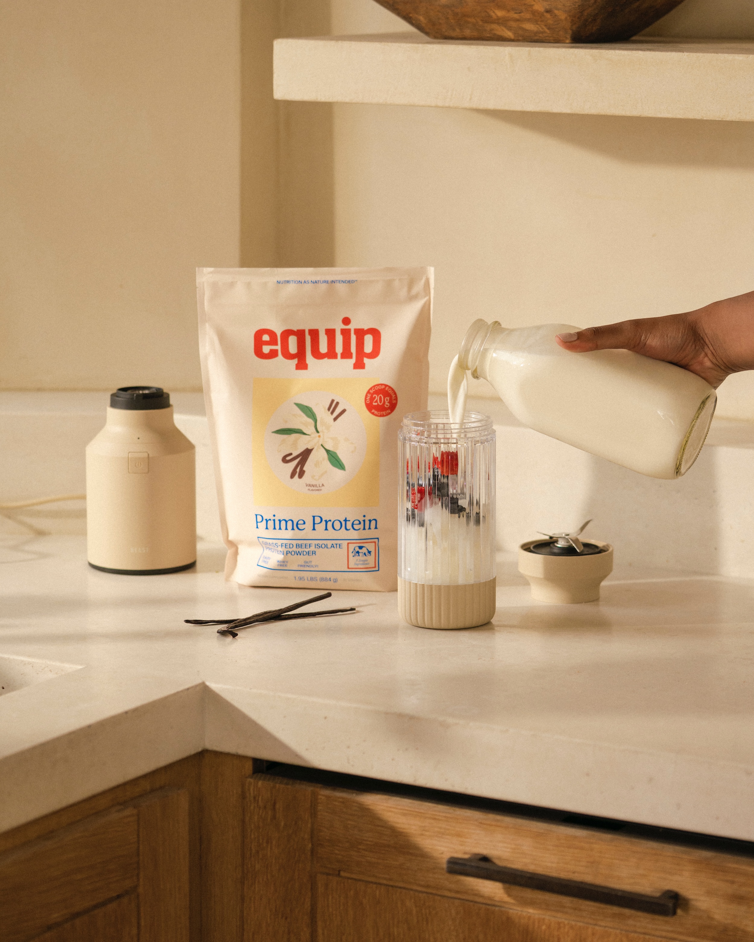 Protein powder by "Equip" in a neutral kitchen on a countertop. Milk pouring into blender.